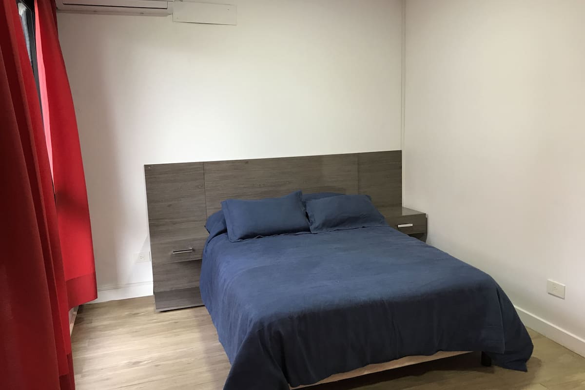 A bedroom with a bed, a nightstand, a drawer, and a red curtain.