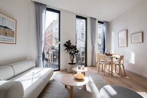 A modern living room with a sofa, a dining table with chairs, a potted plant, and large windows with curtains. - Hausd - Oxford Street (London)