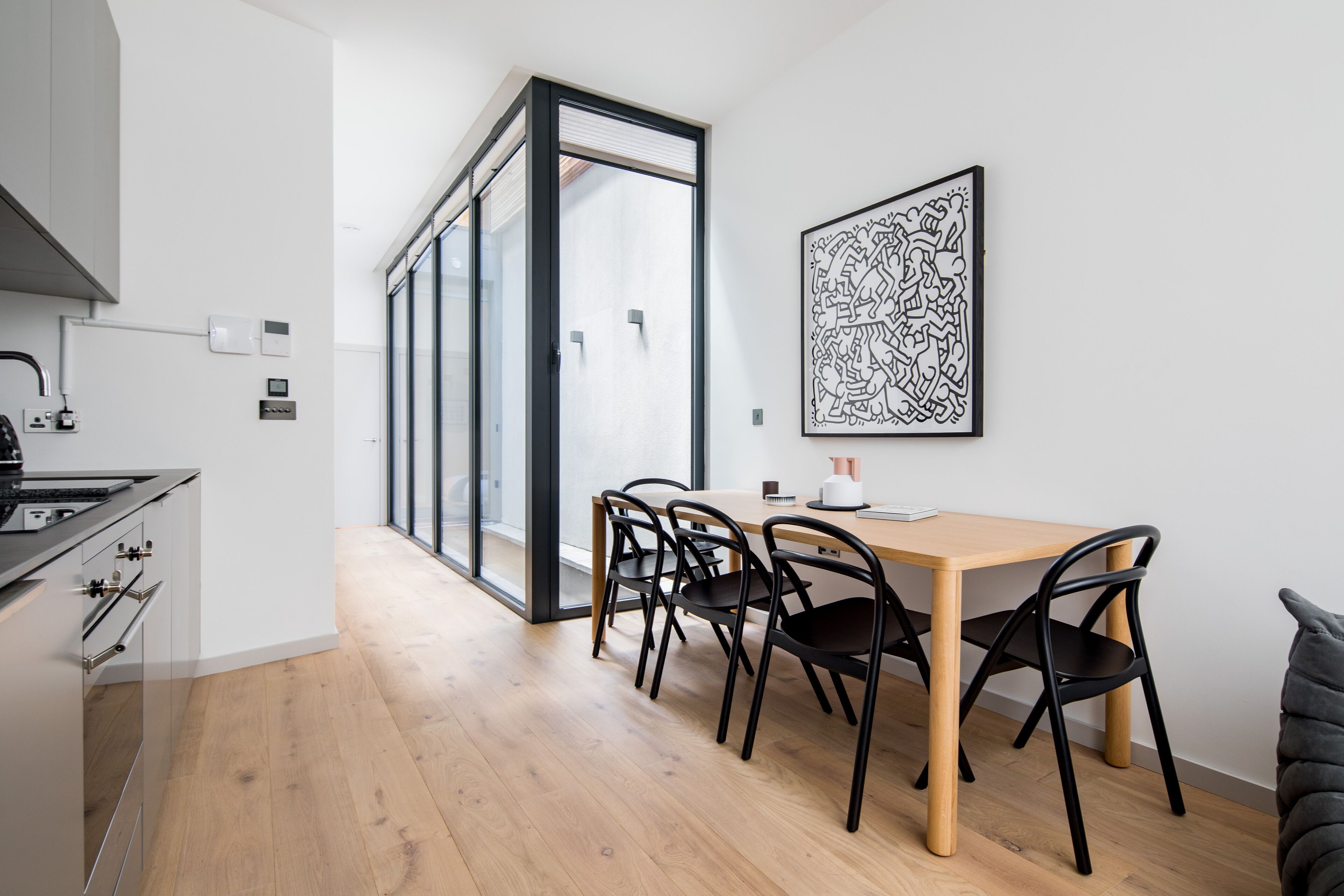 A modern kitchen with a wooden table and chairs, a large window, and a black abstract artwork on the wall.