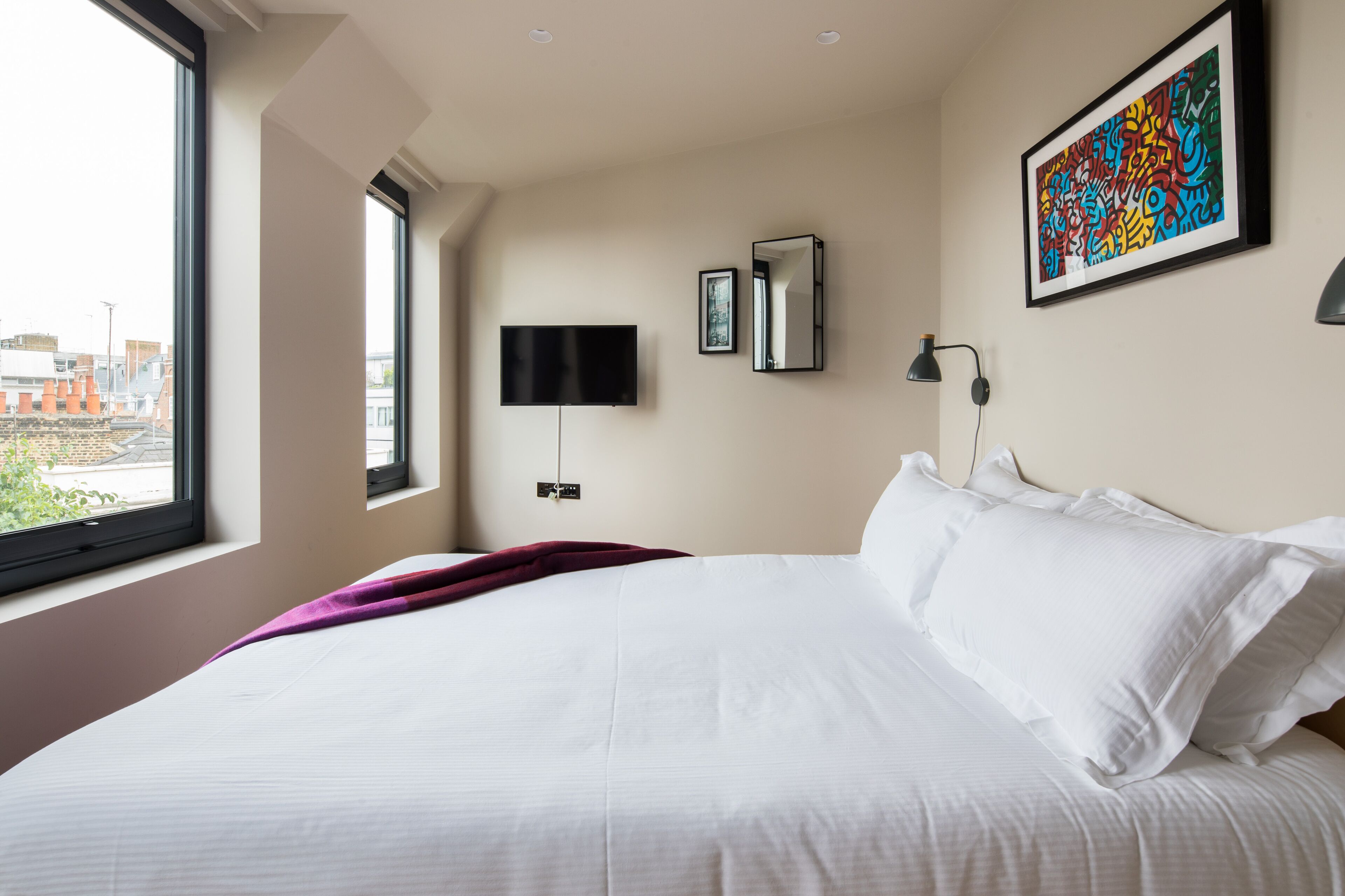 Ein Hotelzimmer mit einem großen Bett, einem an der Wand befestigten Fernseher, einem Fenster mit Blick auf Gebäude und einem farbenfrohen, abstrakten Gemälde an der Wand.