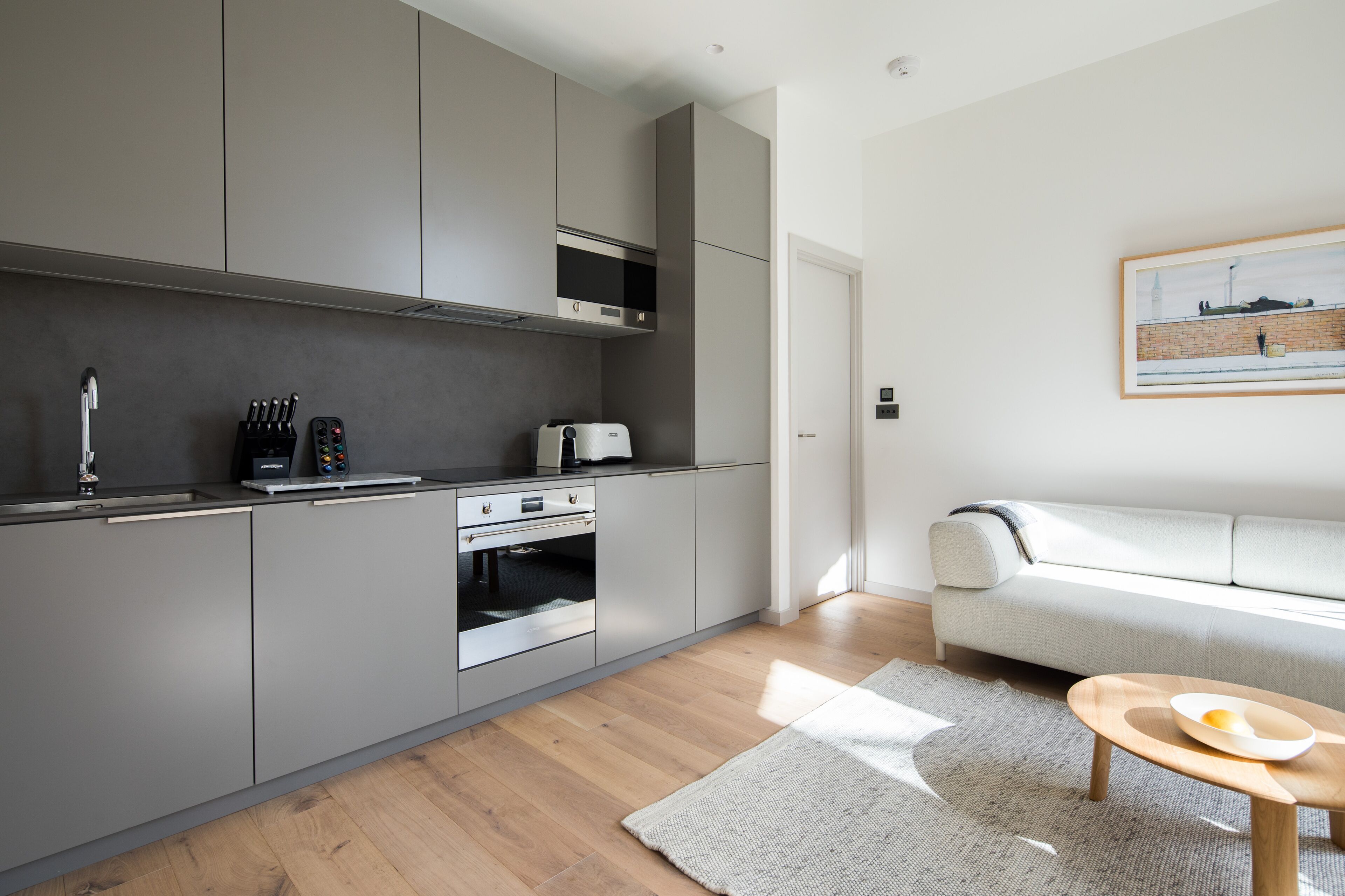Una cucina moderna con mobili grigi, forno integrato e tostapane. C'è un tavolo di legno con una ciotola e un divano bianco nella zona living.