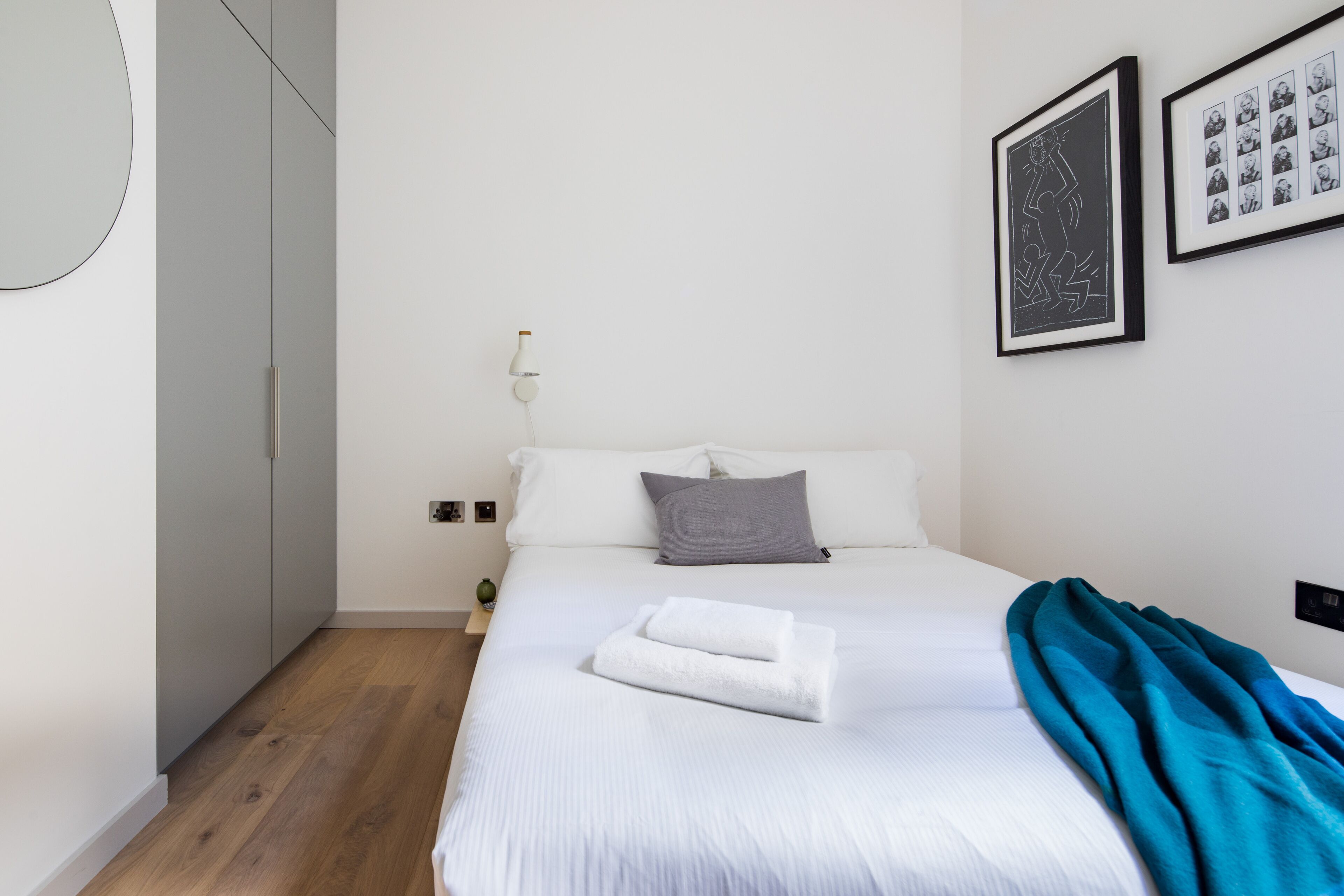 Un letto rifatto con lenzuola bianche e una coperta color petrolio, un cuscino grigio e un quadro appeso al muro.