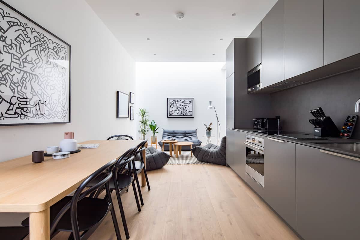 A modern kitchen with grey cabinets, a wooden dining table, and black chairs. There is a large black and white artwork on the wall, a small plant, and a coffee machine.