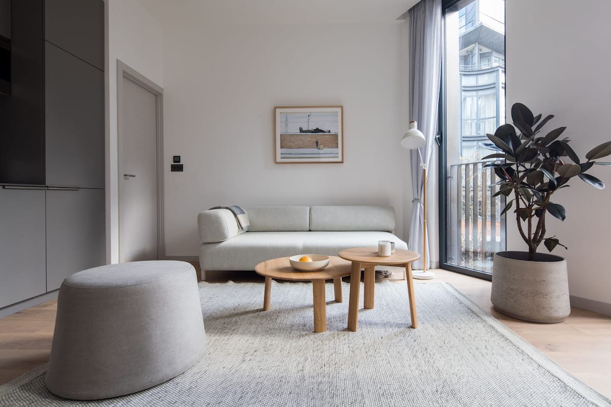 A modern living room with a sofa, ottoman, two small tables, a potted plant, and a framed picture on the wall.