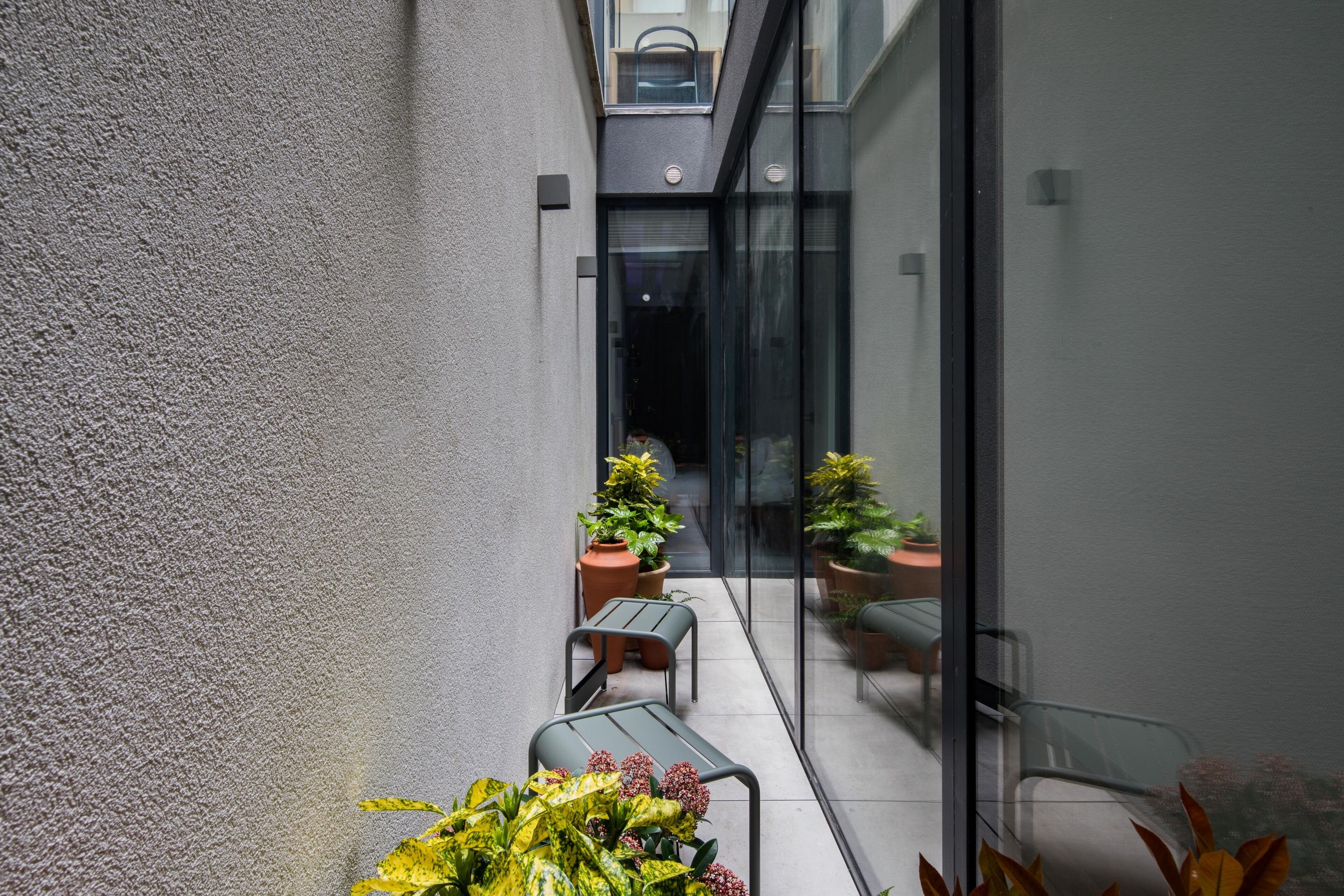 A narrow outdoor corridor with a glass door leading to a balcony. There are potted plants and a small table with chairs along the corridor.