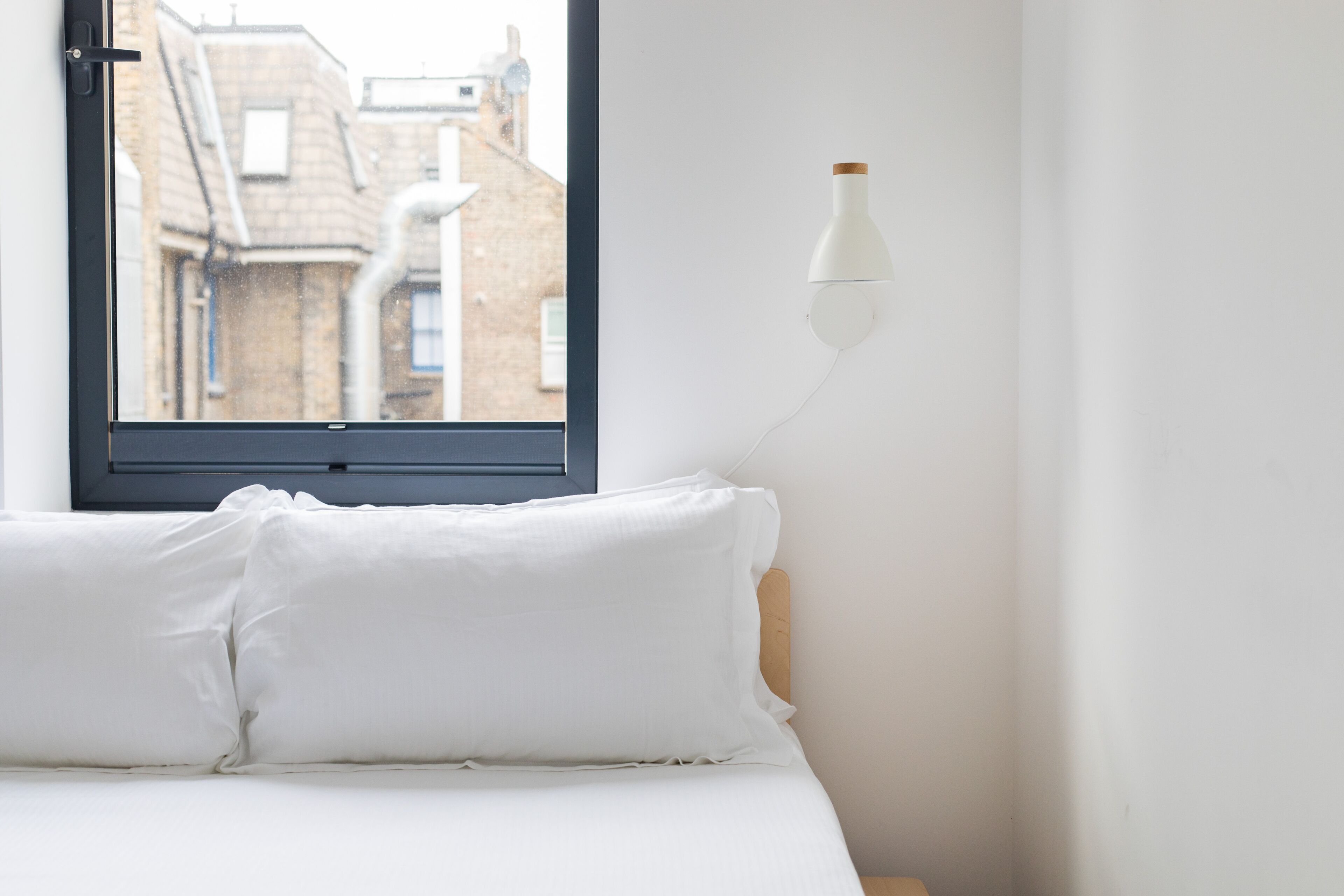 A bedroom with a bed, white pillows, a window with a view of buildings, and a wall-mounted lamp.