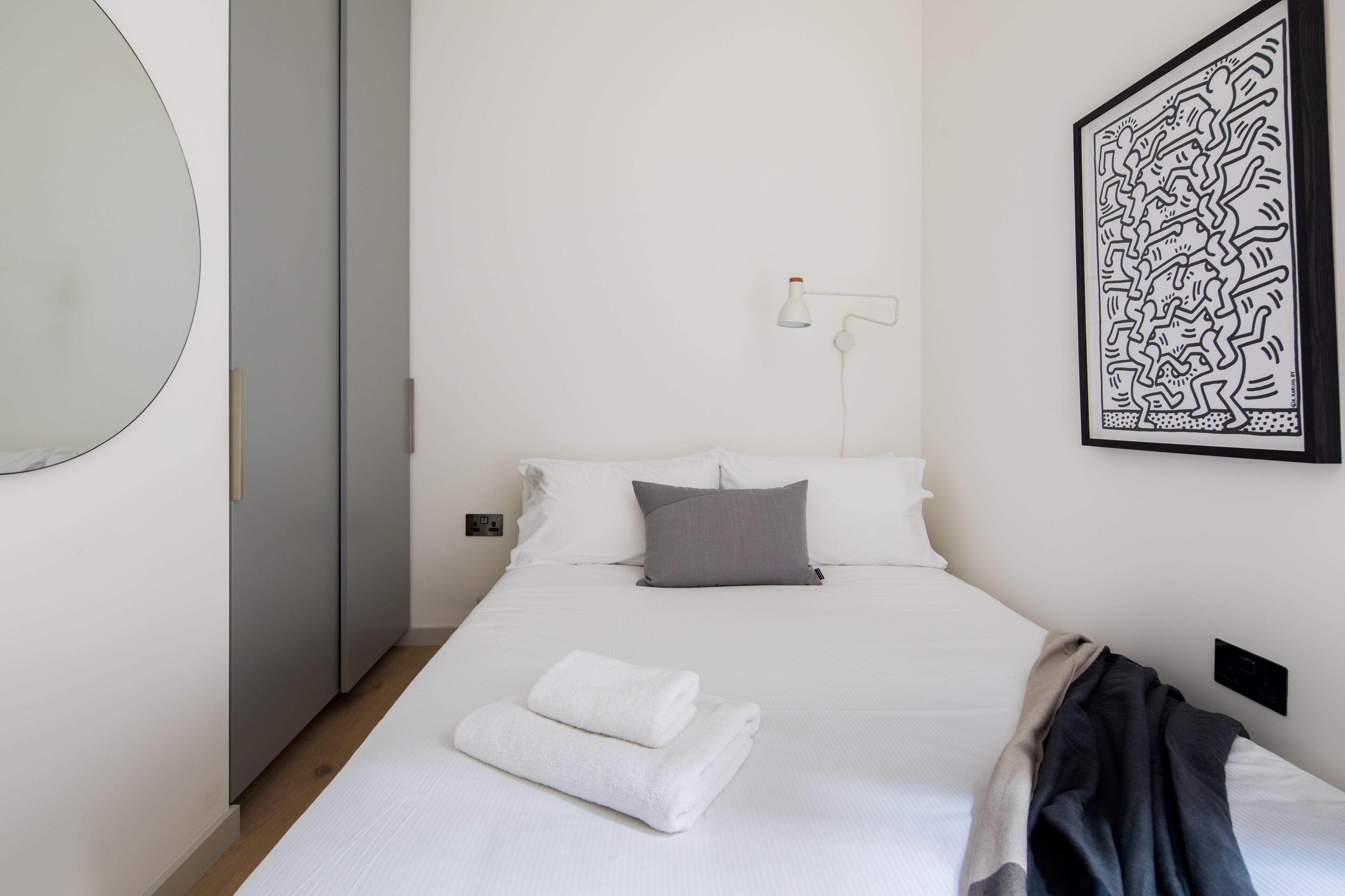 Ein ordentlich bezogenes Bett mit weißen Leinen, einem grauen Kopfkissen und einem gefalteten weißen Handtuch. An der Wand hängt ein Schwarz-Weiß-Kunstwerk und neben dem Bett steht eine weiße Stehlampe.
