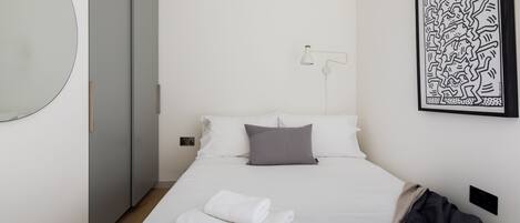 Ein ordentlich bezogenes Bett mit weißen Leinen, einem grauen Kopfkissen und einem gefalteten weißen Handtuch. An der Wand hängt ein Schwarz-Weiß-Kunstwerk und neben dem Bett steht eine weiße Stehlampe.