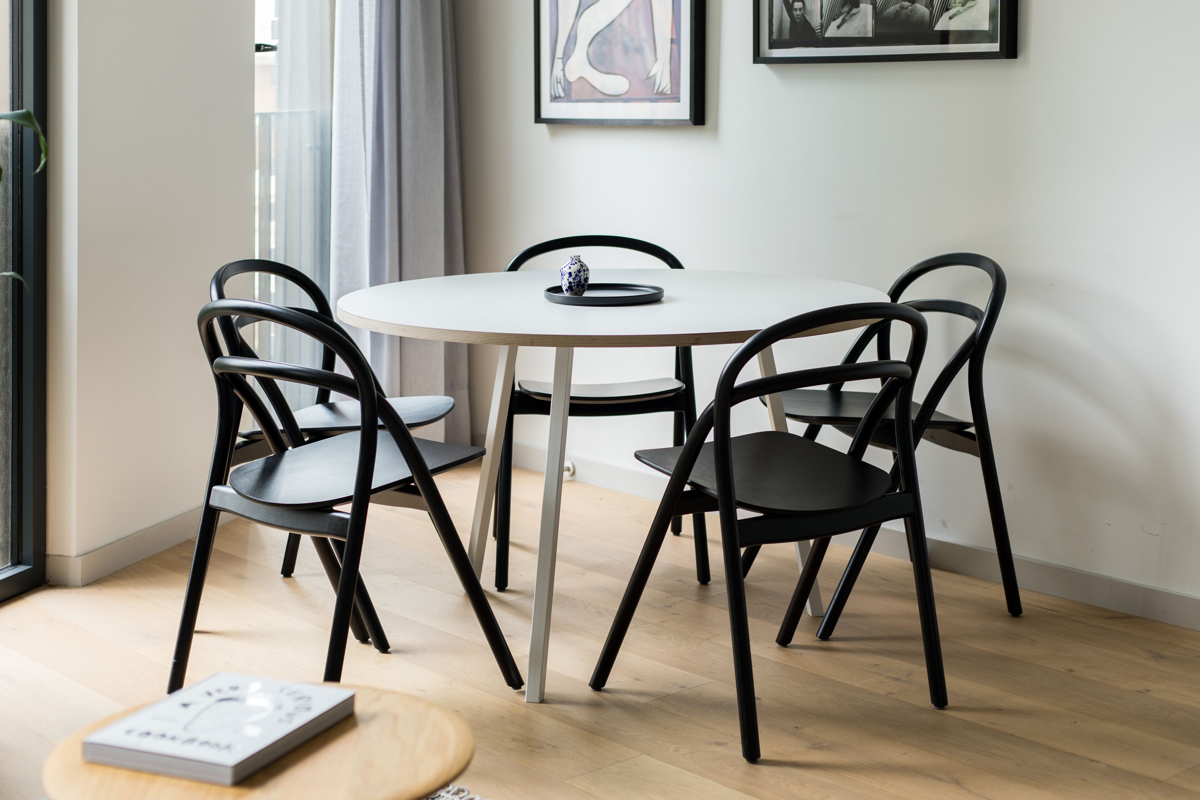 A round dining table with four black chairs, a small white table lamp, and a framed artwork on the wall.