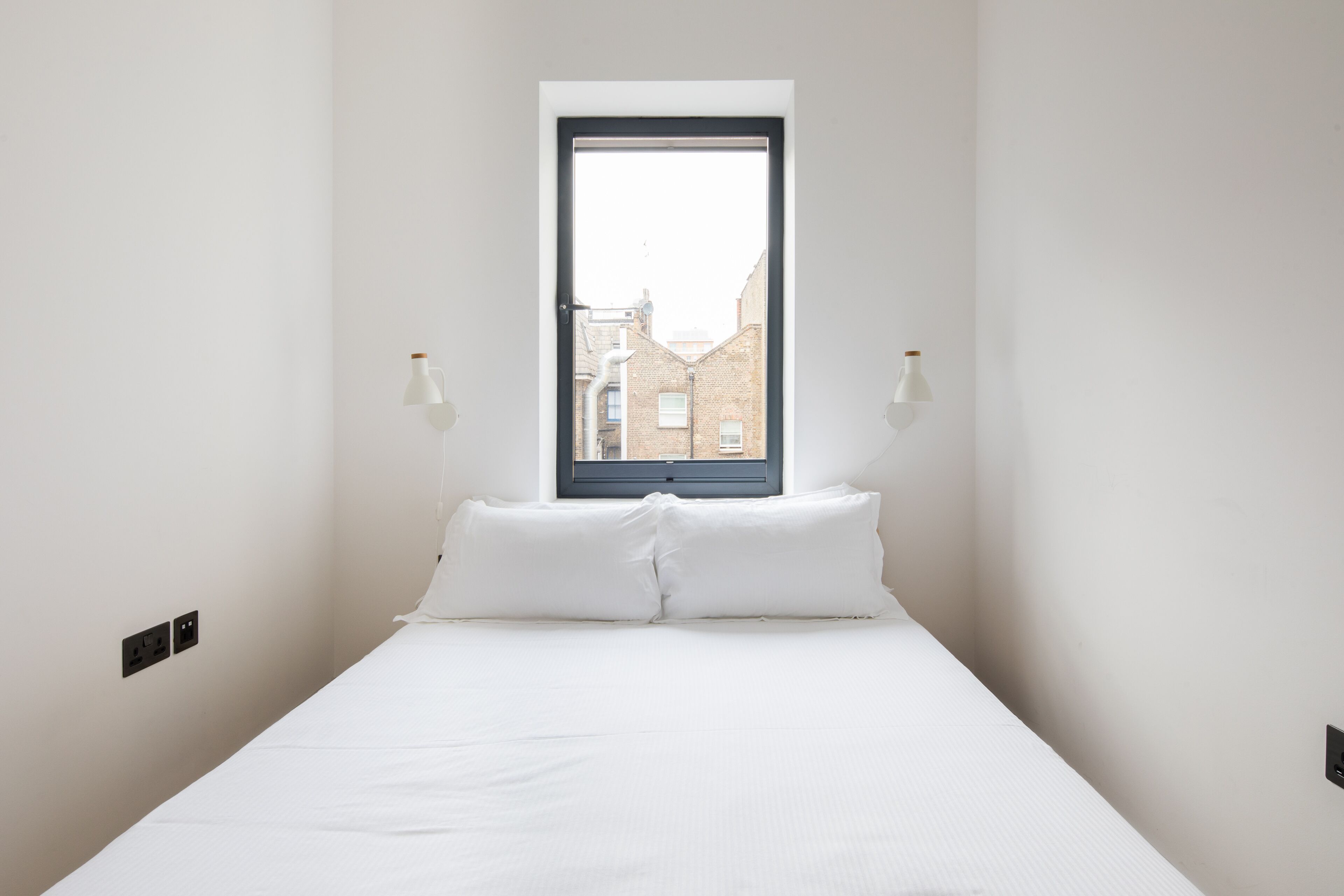 A minimalist bedroom with a single bed, a window, and two wall-mounted lamps.