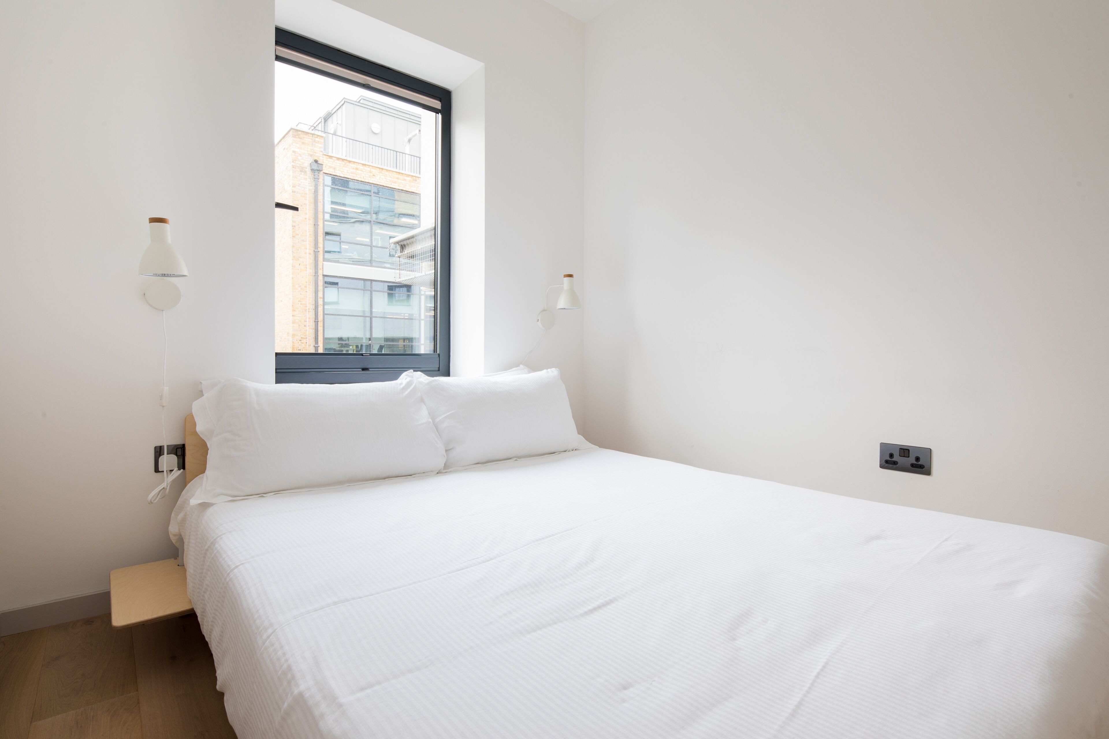 Une chambre à coucher au style minimaliste, avec un grand lit, une fenêtre et deux lampes fixées au mur.