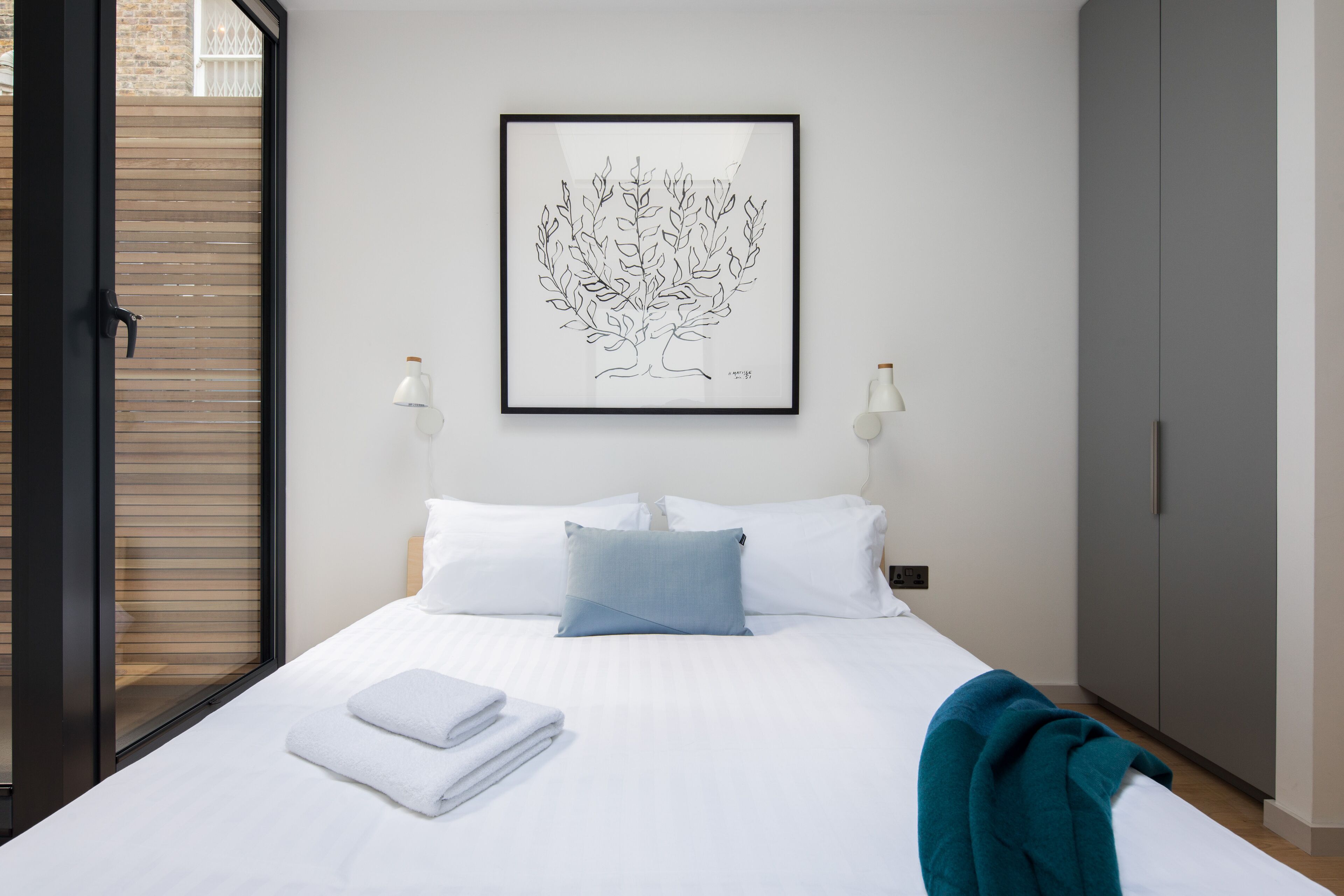 A neatly made bed with white linens, a blue pillow, and a teal throw blanket. There is a framed artwork of a tree on the wall above the bed.