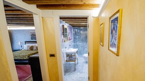 A narrow hallway with wooden flooring, a bed with a patterned bedspread, a bathroom with a sink and mirror, and a framed picture on the wall. - EGO' Residence Venice (Venice)