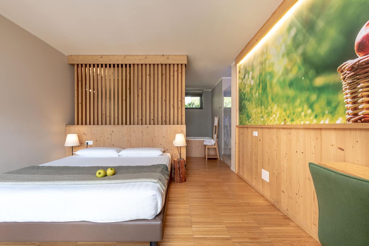 A bedroom with a wooden headboard, a bed with white linens, two apples on the bed, a green chair, and a wall with a nature-themed artwork.