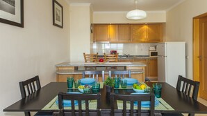 Un comedor con una mesa preparada para cuatro, una cocina con armarios de madera y un refrigerador, y cuadros en la pared.