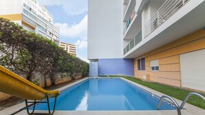 Piscina con un tobogán amarillo, rodeada de follaje y edificios modernos.