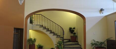 Un interior amplio con una escalera central, varias plantas en macetas y un piso de baldosas.