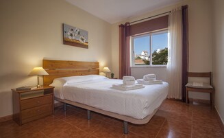 A hotel room with a bed, two bedside lamps, a nightstand, a chair, a window with curtains, and a painting on the wall.
