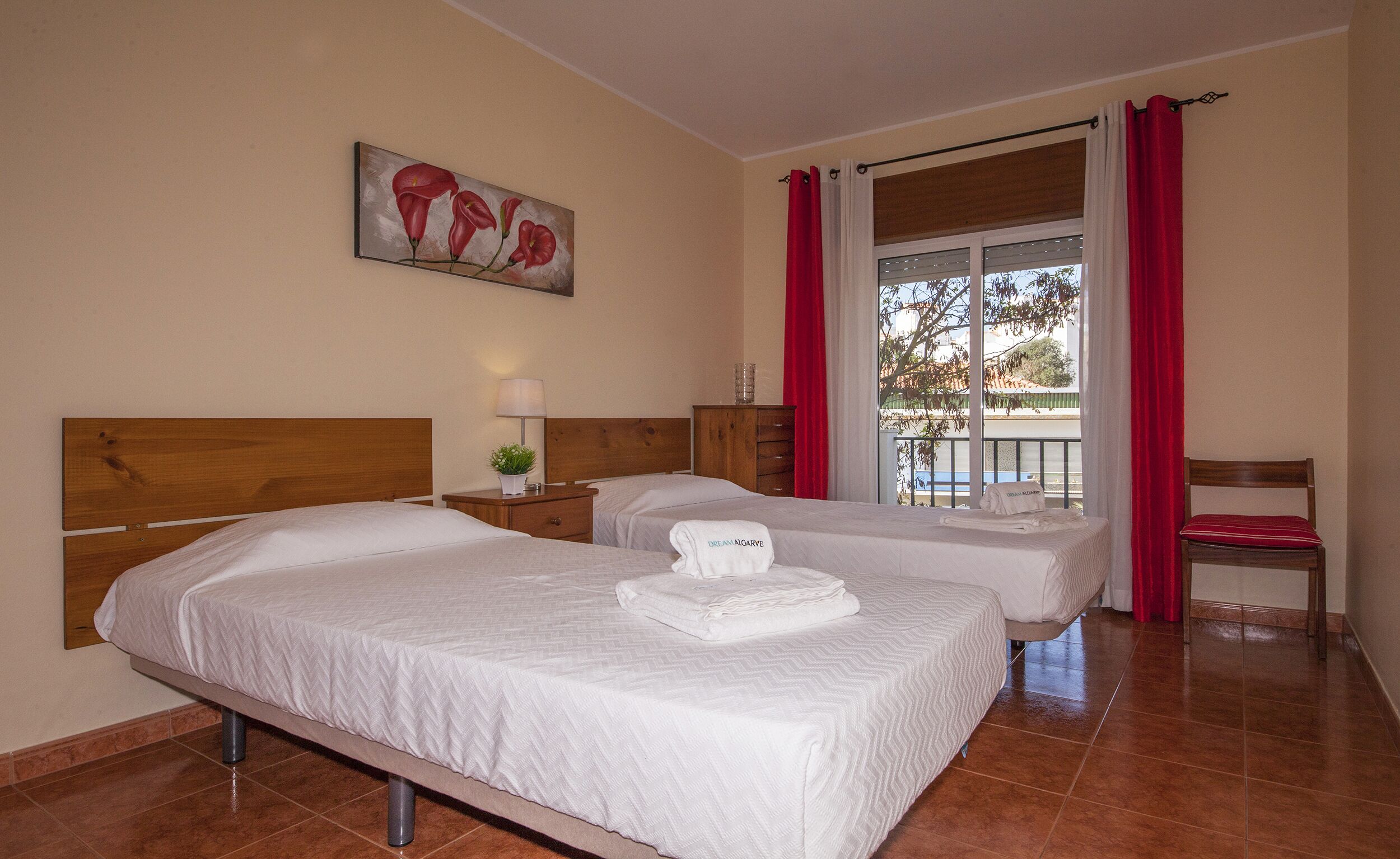 A hotel room with two beds, a painting on the wall, a window with red curtains, and a chair.