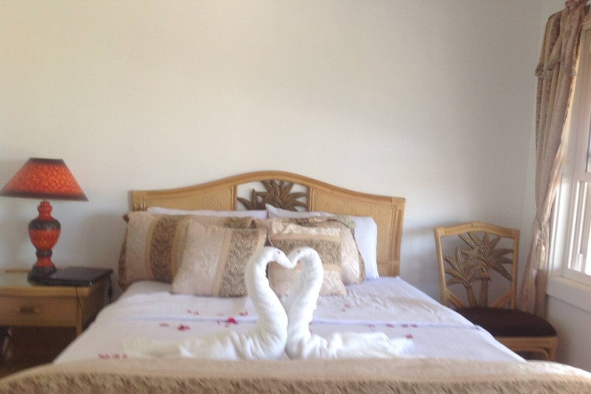 A neatly made bed with a heart-shaped towel arrangement, a wooden headboard, a nightstand with a lamp, and a chair with a palm leaf design.