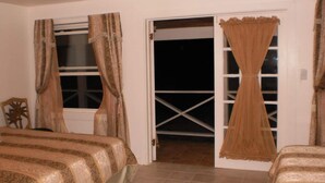 Un dormitorio con dos camas, un ventanal con cortinas, una puerta que da a un balcón y un ventilador de techo.
