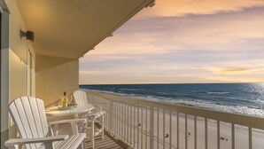 A balcony with white chairs and a table, overlooking a beach with the ocean and a sunset.