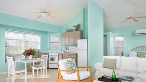 A bright living room with a white sofa, a wooden chair, a dining table with chairs, a kitchen area with cabinets and appliances, and a ceiling fan. - Cottage for 2 at Dragon Cay Resort Mudin Harbour (Mudjin Harbor)