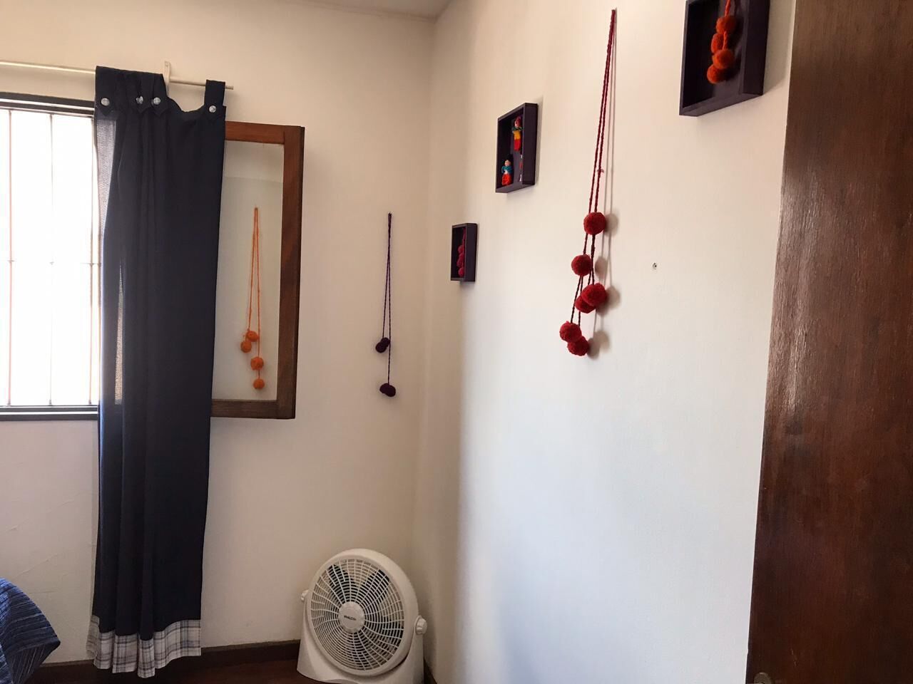 A room with a bed, a standing fan, a window with a curtain, and wall decorations including framed pictures and hanging ornaments.