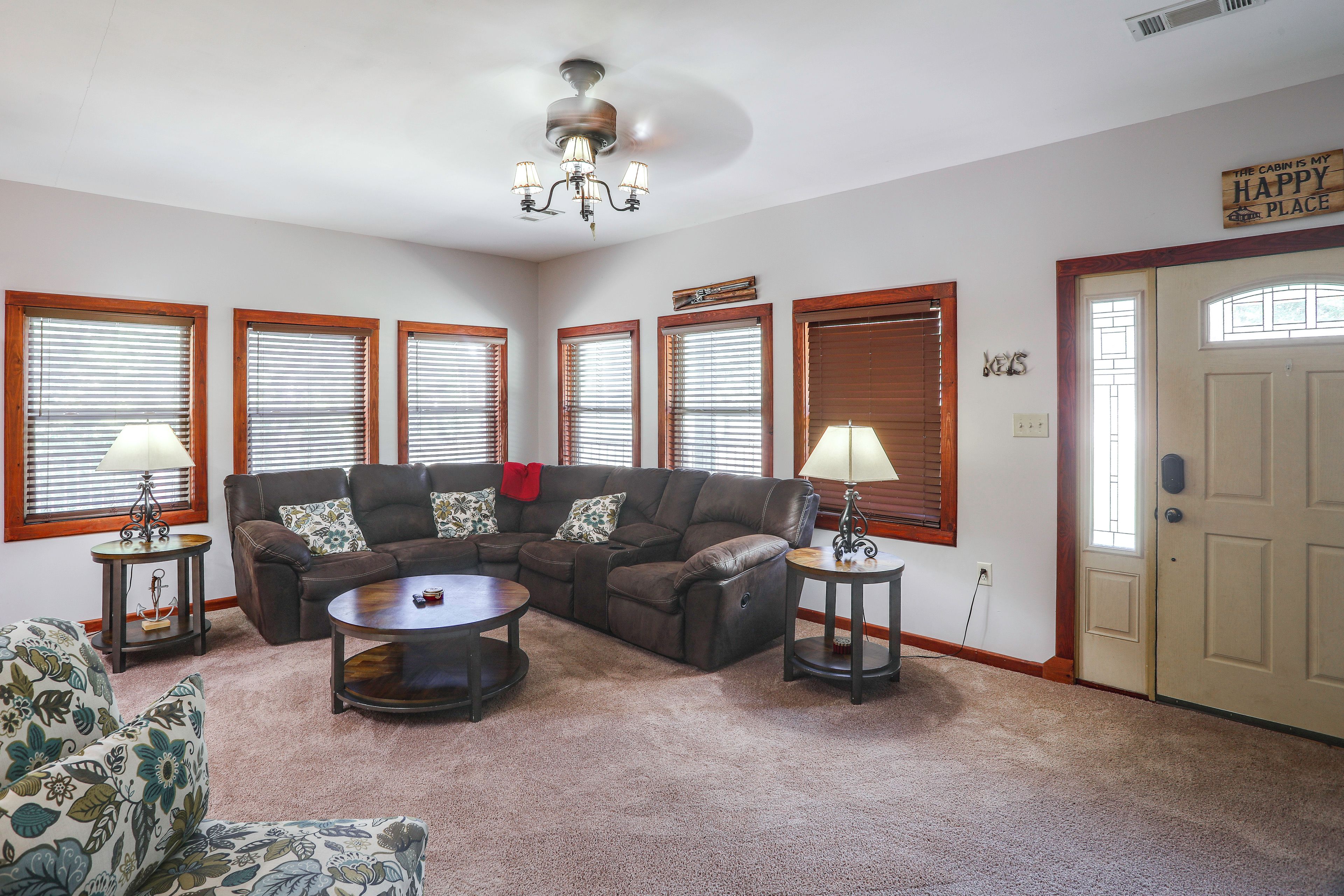 A living room with a brown leather sofa, a round coffee table, and a chandelier. There are four windows with blinds, a door, and a wall sign that says 'The Cabin is My Happy Place.'