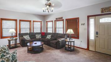 A living room with a brown leather sofa, a round coffee table, and a chandelier. There are four windows with blinds, a door, and a wall sign that says 'The Cabin is My Happy Place.'