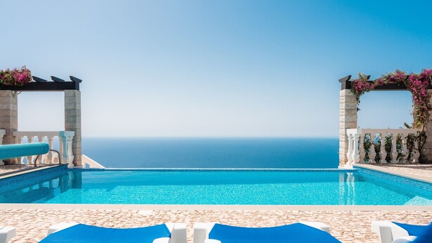 Une piscine offrant une vue dégagée sur l’océan, entourée de murs en pierre et de chaises longues bleues.