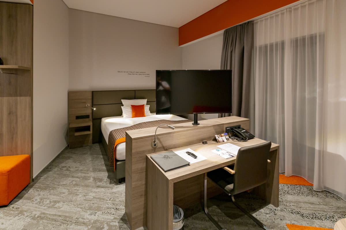 Habitación de hotel con cama, escritorio, computadora, televisor y ventana con cortinas.