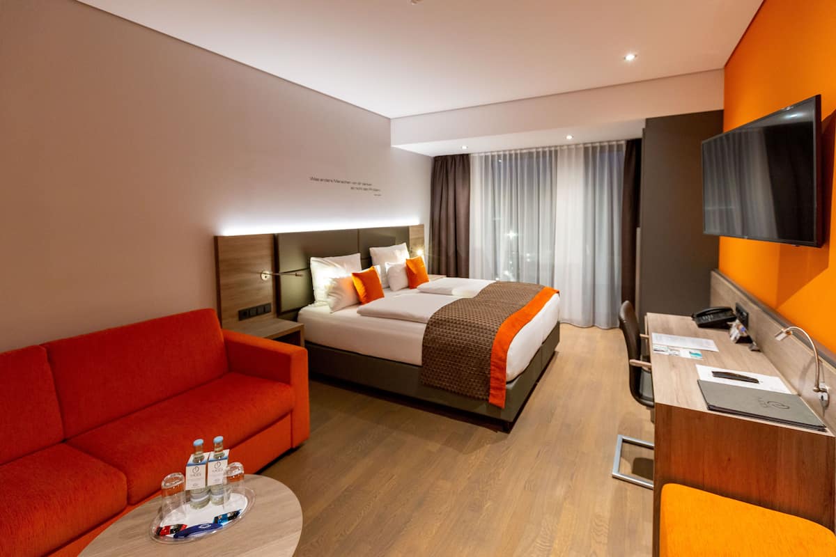 Habitación de hotel con cama, sofá rojo, escritorio con silla, televisión y ventana con cortinas.