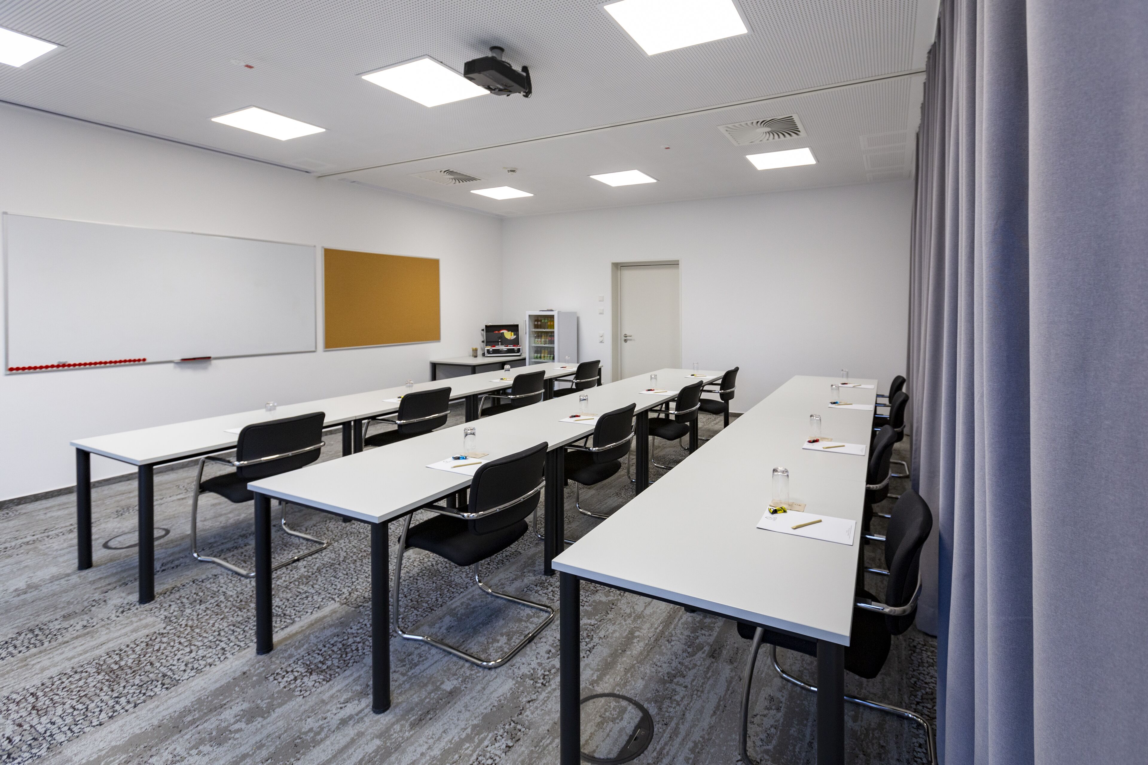 Un salón de conferencias con mesas largas, sillas, una pizarra y un proyector.