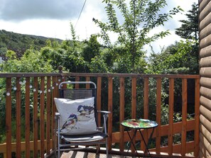 A wooden deck with a metal chair and a small table, surrounded by lush greenery and a mountainous backdrop. - 2 bedroom accommodation in Creetown, near Newton Stewart (Creetown, near Newton Stewart)