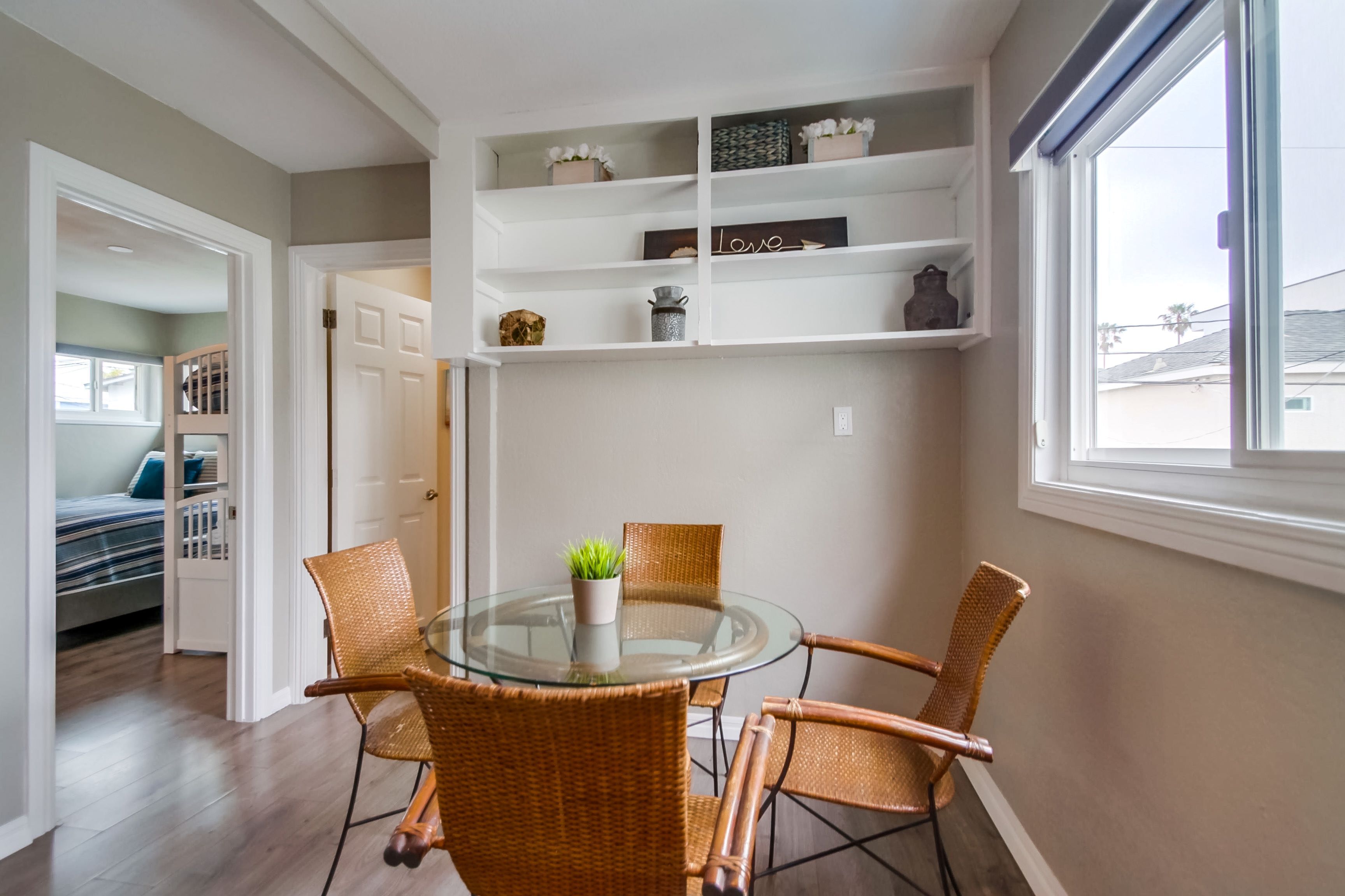 A dining area with a glass table and wicker chairs, a white shelving unit above, and a view through a window.