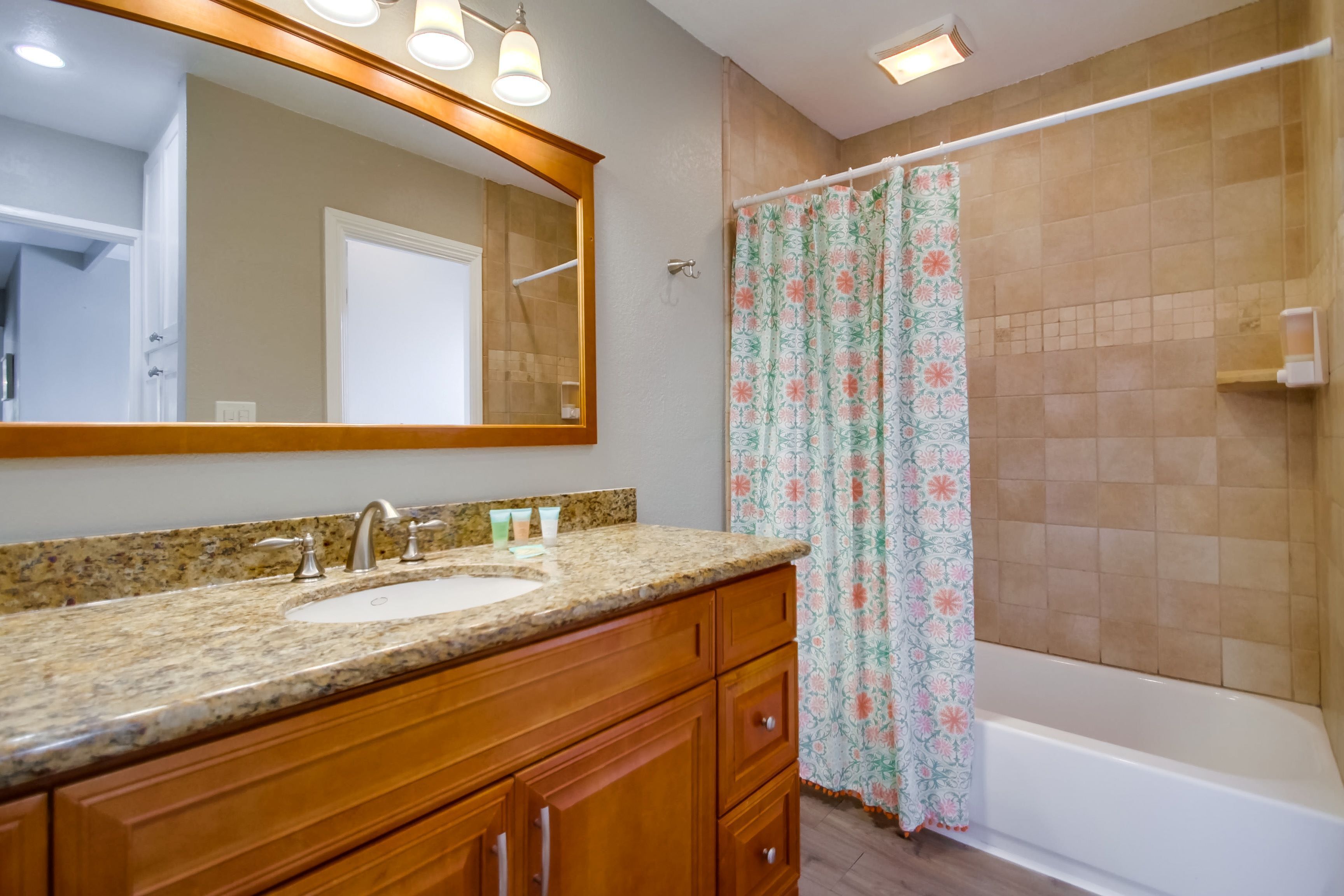 A bathroom with a granite countertop sink, wooden cabinets, a large mirror, and a shower with a floral curtain.
