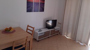 A small, tidy room with a wooden dining table, a single chair, a TV on a white cabinet, and a wall-mounted picture of lavender fields.