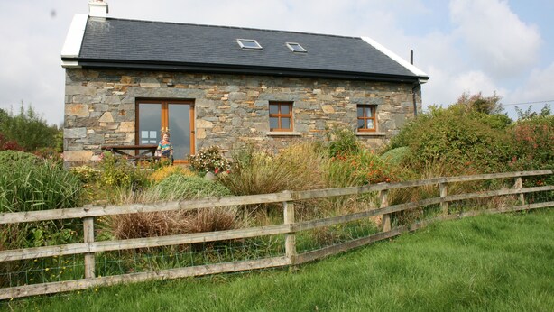 A stone house with a sloped roof, two windows, and a wooden fence in the foreground. - Groff Cottage (Tourmakeady)