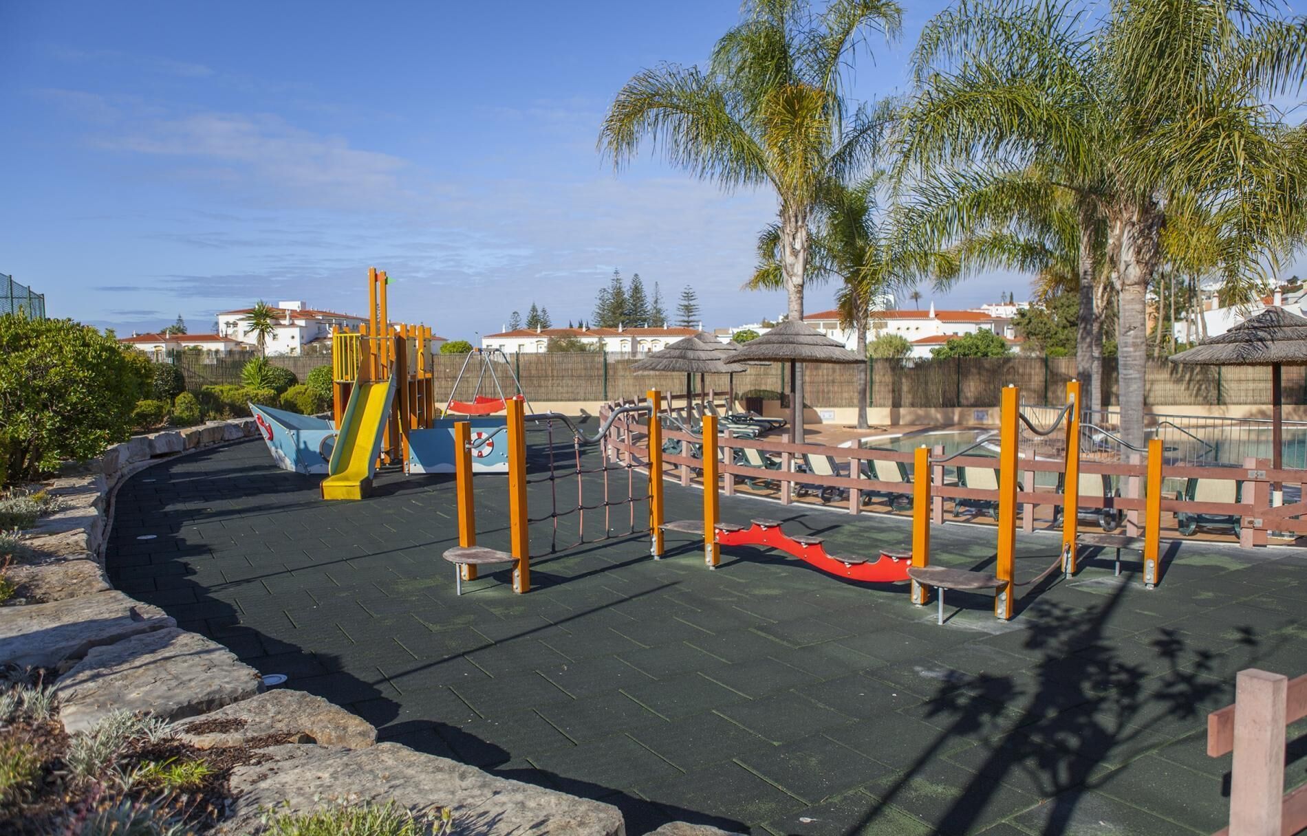 A playground with various play structures, including slides and climbing frames, surrounded by a paved area and palm trees.