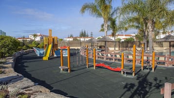 A playground with various play structures, including slides and climbing frames, surrounded by a paved area and palm trees.