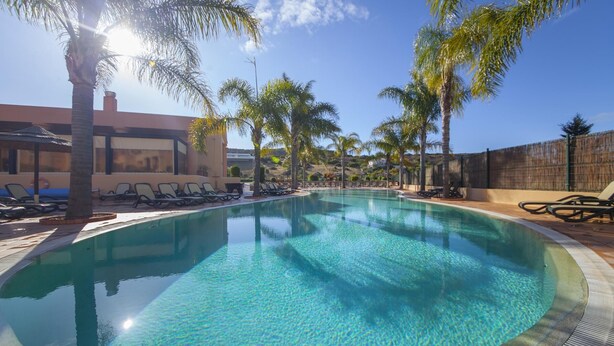 A clear swimming pool surrounded by palm trees and lounge chairs, with a building in the background.
