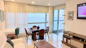 Una sala de estar moderna con sofá, mesa de centro, comedor y una pantalla de televisión mostrando el precio de una habitación.