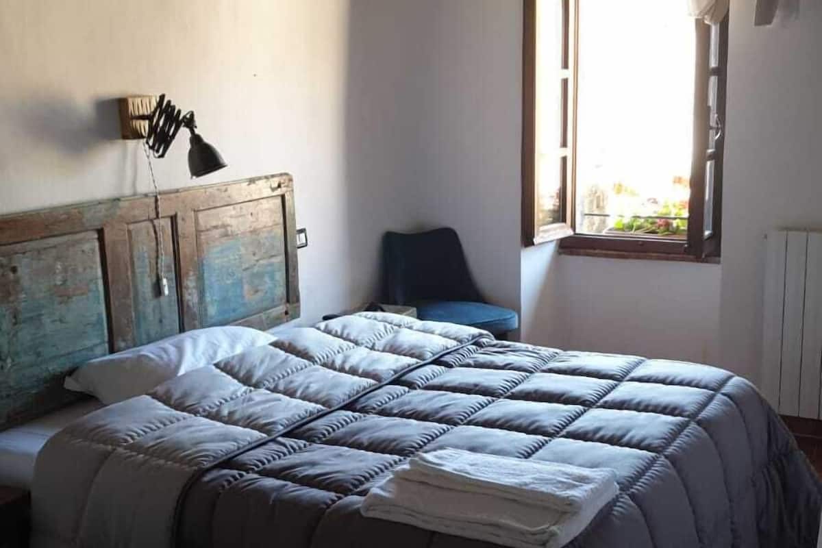 A bedroom with a large bed, a smaller bed, a chair, a wooden headboard, a window with curtains, and a radiator.