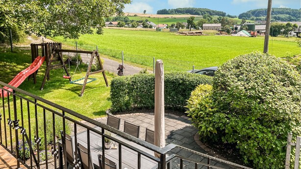 Een speeltuin met een glijbaan en schommels, omringd door een verzorgd gazon en een uitzicht op een landelijk landschap met velden en gebouwen in de verte.