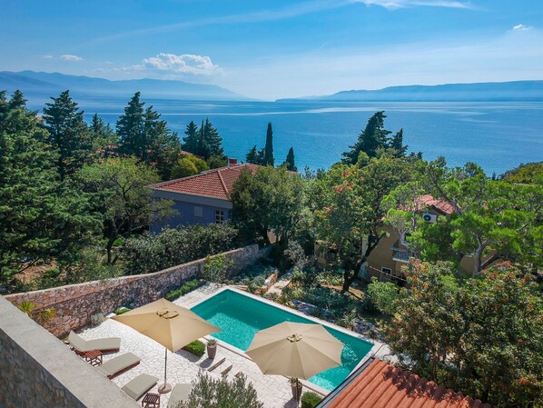 A pool area with two umbrellas, a stone wall, and a view of the sea and mountains.