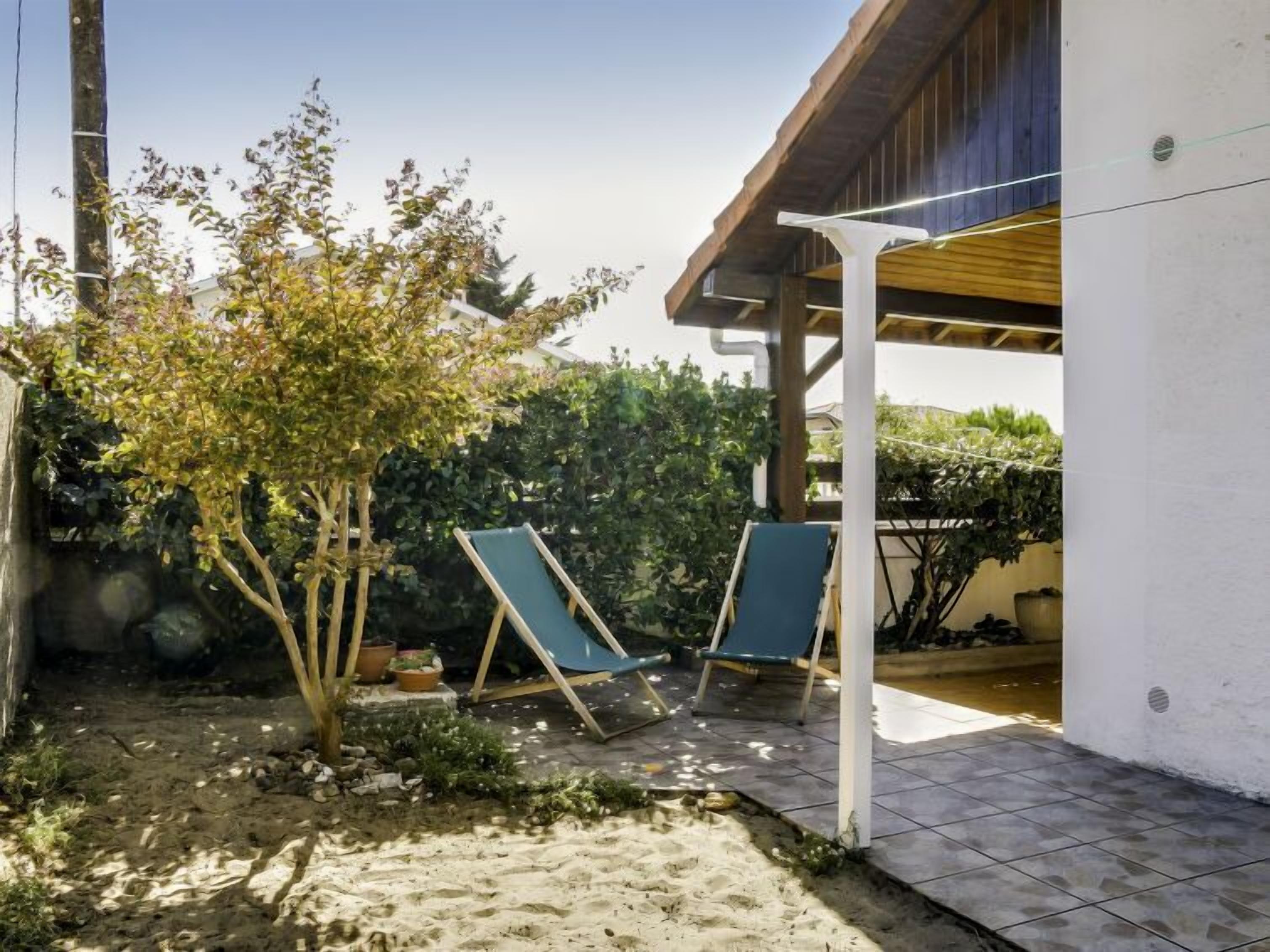A covered outdoor area with two lounge chairs, a small tree, and a white building.