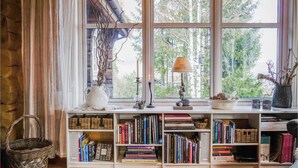 Un coin lecture confortable avec une grande fenêtre, des étagères remplies de livres, un vase décoratif et une bougie posée sur le rebord d’une fenêtre.