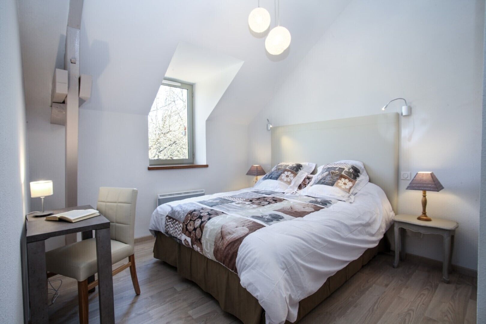 Ett sovrum med en stor säng, två sänglampor, ett litet bord med en stol och ett fönster med utsikt över träd.