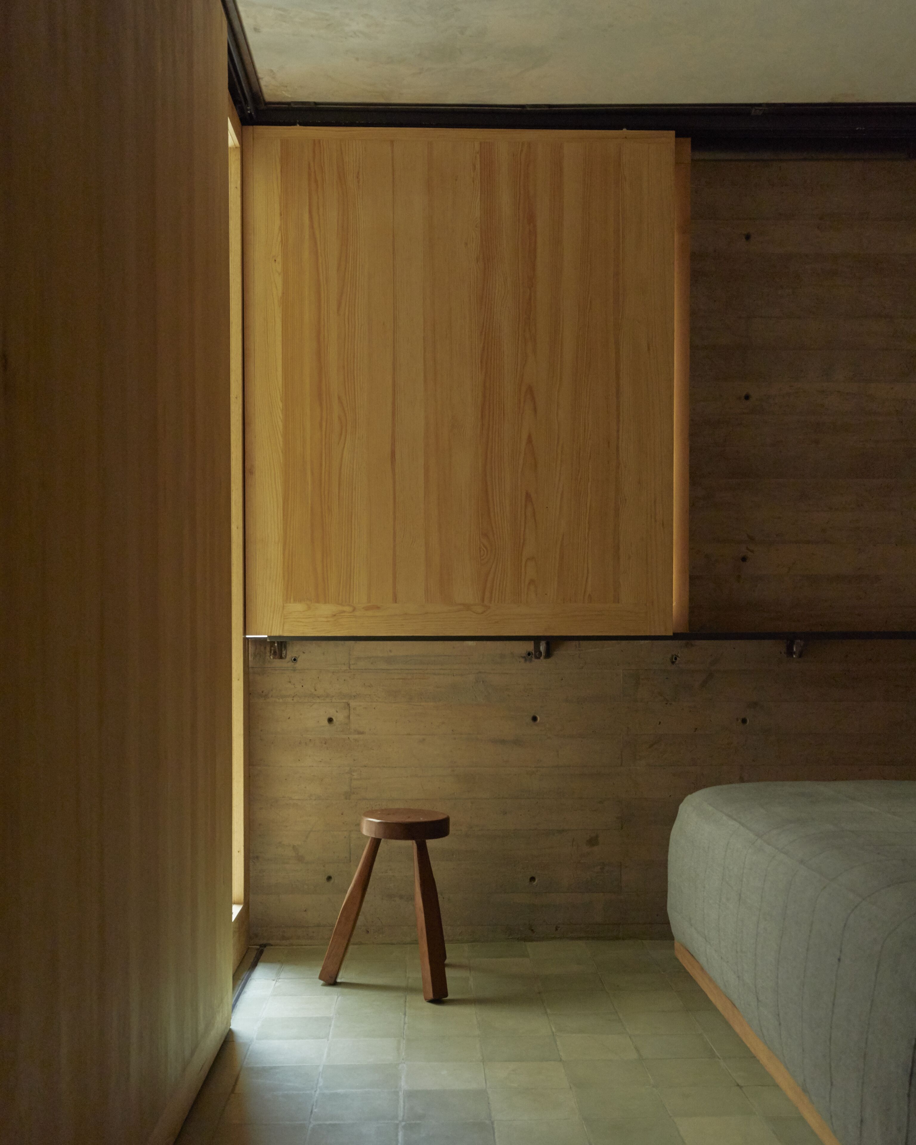 A room with a wooden wardrobe, a small stool, and a bed.